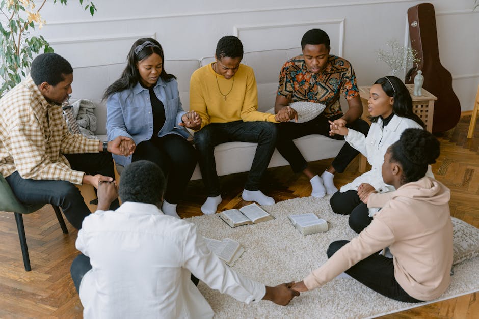A diverse group of young adults sitting in a circle, holding hands and praying together with open Bibles on the floor in a living room setting.